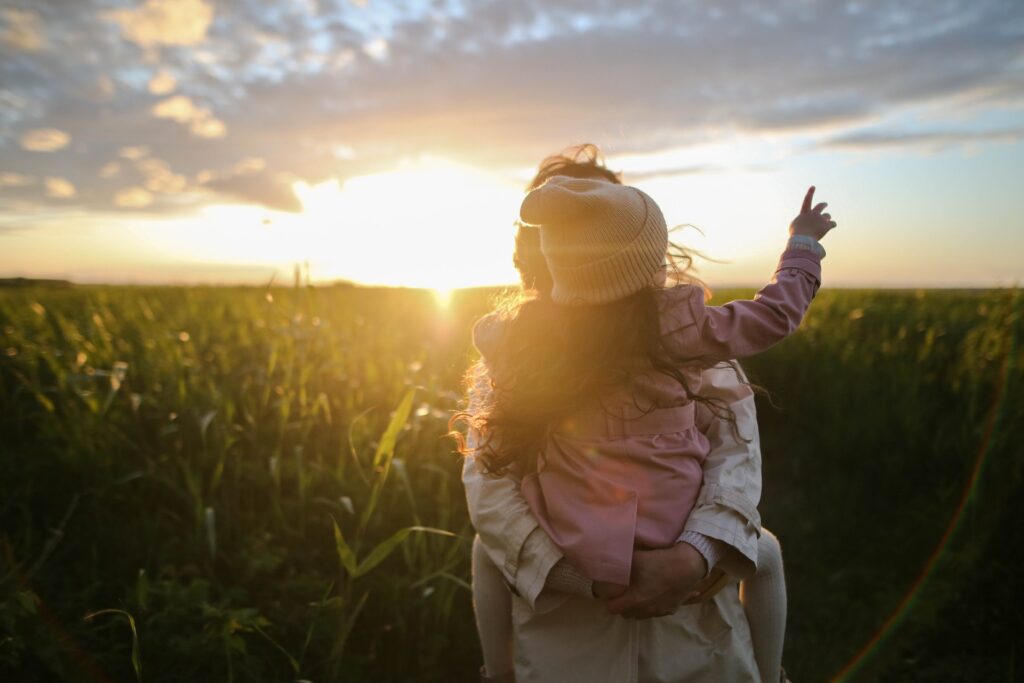 A Mom pointing in the air carrying a child with a serene stress free field overlooked by a beautiful sunset