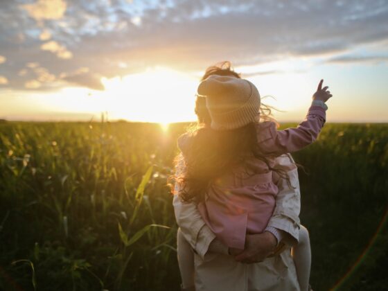 A Mom pointing in the air carrying a child with a serene stress free field overlooked by a beautiful sunset
